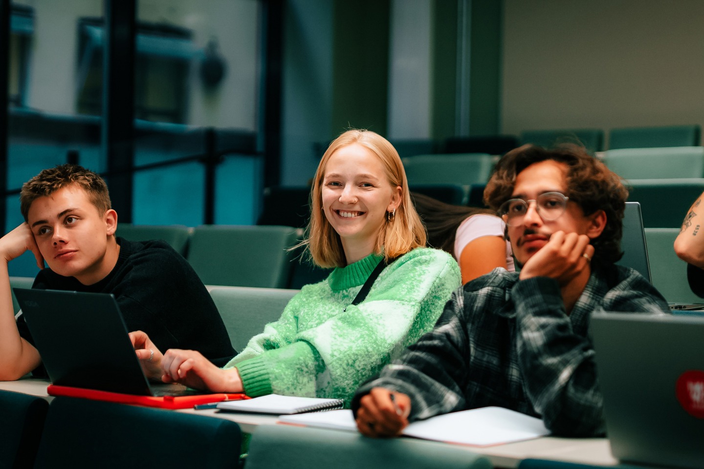 Bildet viser tre unge som sitter i en forelesningssalen. Kvinnen på bildet smiler og de to unge mennene på hver side ser interessert på tavlen.