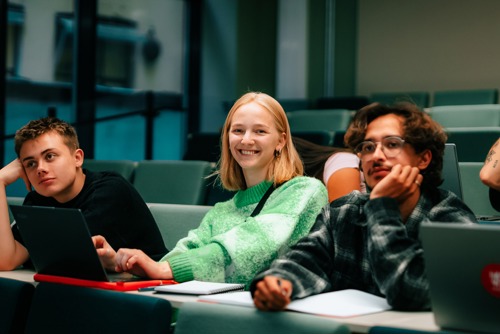 Bildet viser tre unge som sitter i en forelesningssalen. Kvinnen på bildet smiler og de to unge mennene på hver side ser interessert på tavlen.