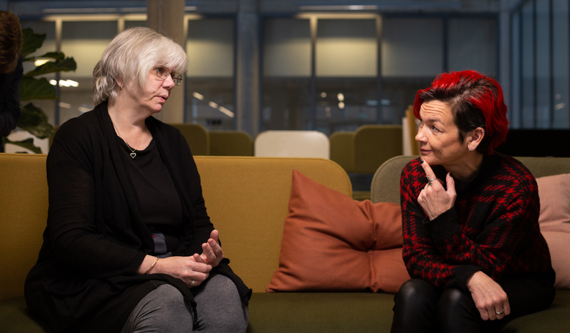 Faglærer i helsekoordinatorstudiet Marit Berg sitter i en sofa og snakker med Manuela Rothe. Marit har grått hår og svart cardigan, og Manuela har rødt, kort hår, og rød rutete genser, og hun har fingeren på haken.