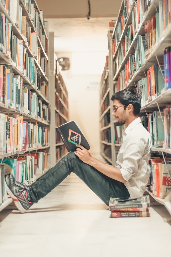 Ung mann sitter på gulvet og leser i en bok på et bibliotek.