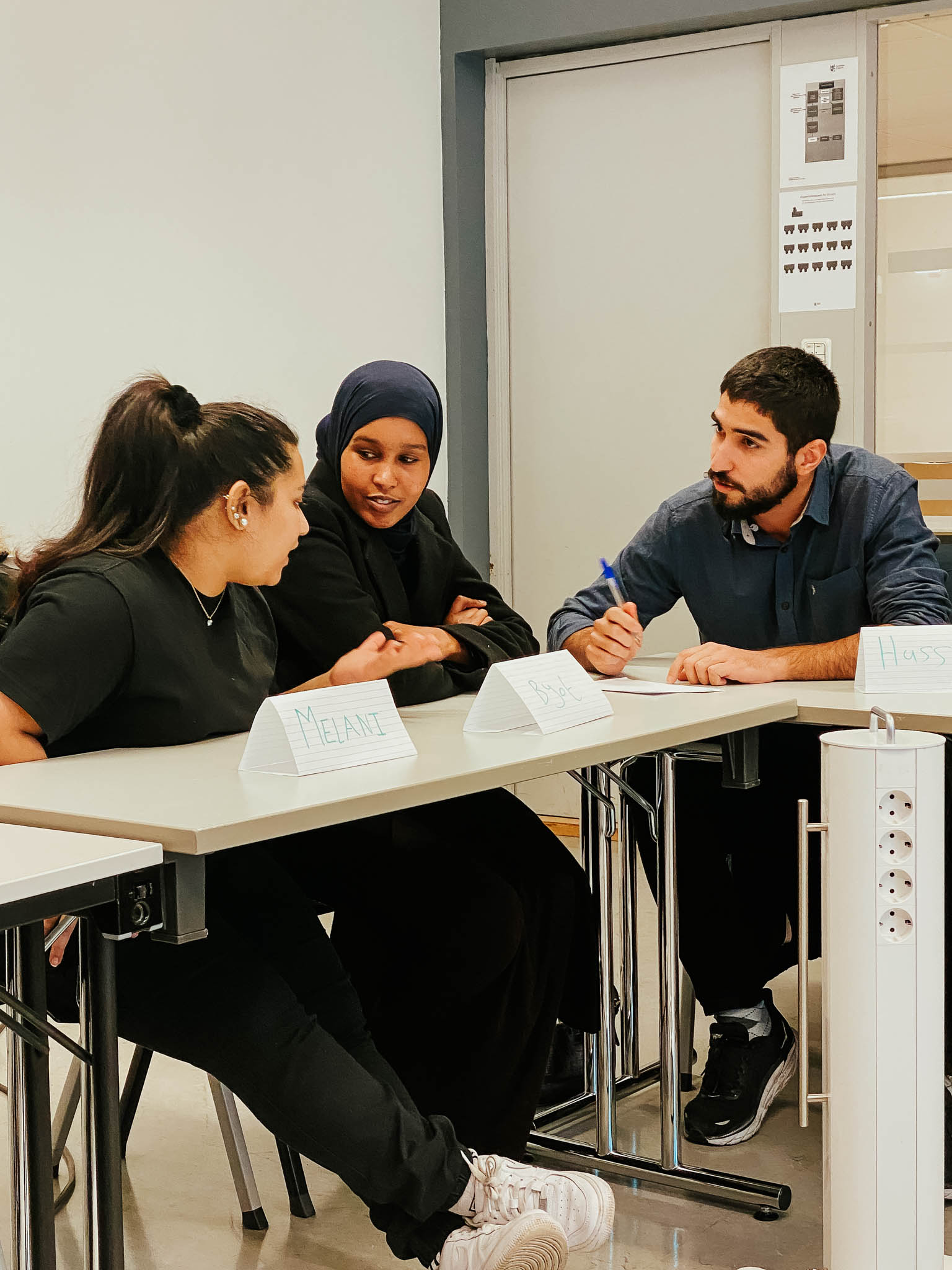 Tre studenter sitter ved en skrivepult mens de diskuterer seg i mellom. Det er en mann og to kvinner som samhandler. Foto.