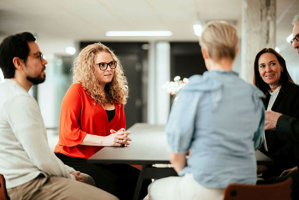En gruppe mennesker i arbeidsmiljø sitter rundt et bord. En kvinne med krøllete hår og en oransje bluse ser oppmerksomt på en kollega, mens resten av gruppen deltar i diskusjonen.