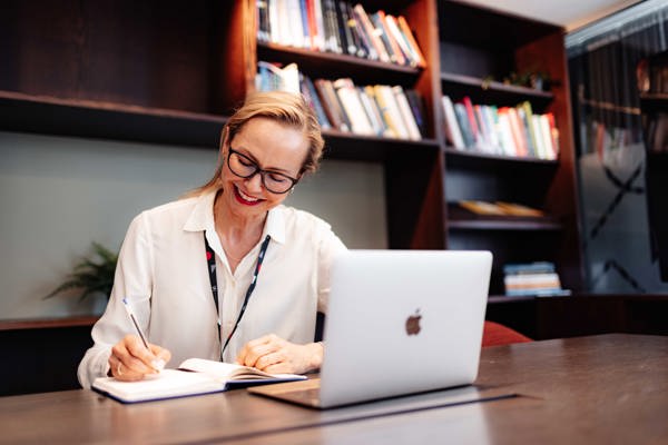En person med briller og en hvit bluse smiler mens de skriver i en notatbok ved et skrivebord med en bærbar datamaskin foran seg, i et kontormiljø med en bokhylle i bakgrunnen.