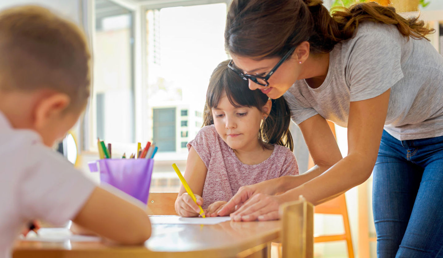 En voksen hjelper et barn med å tegne ved et bord i et lyst klasserom, mens et annet barn fokuserer på sitt eget arbeid i forgrunnen.