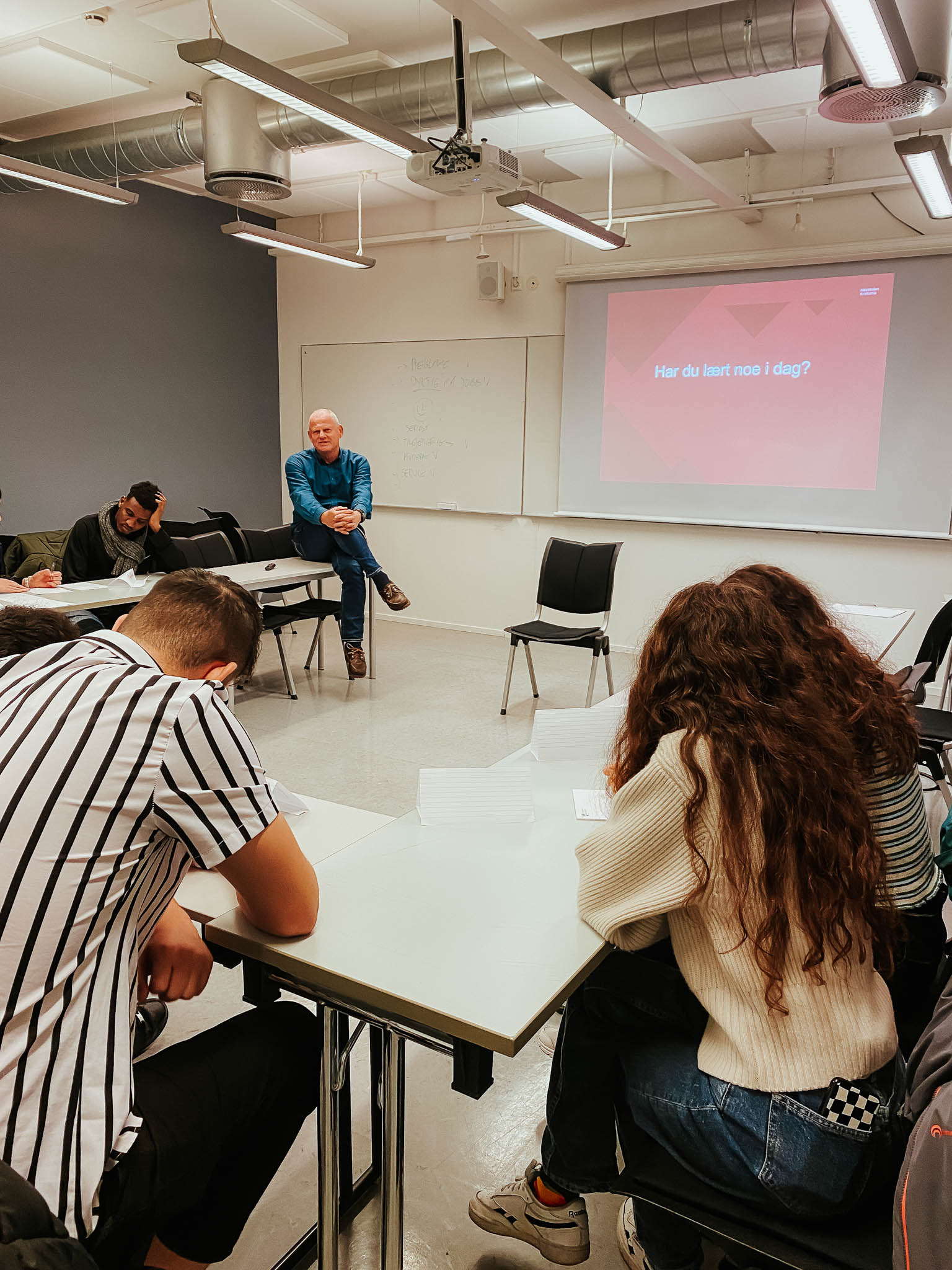 Studentene sitter med bøyde nakker over skrivepulter i en firkantformasjon vendt mot professor Nils Høgevold som foreleser til forsamlingen. Foto.