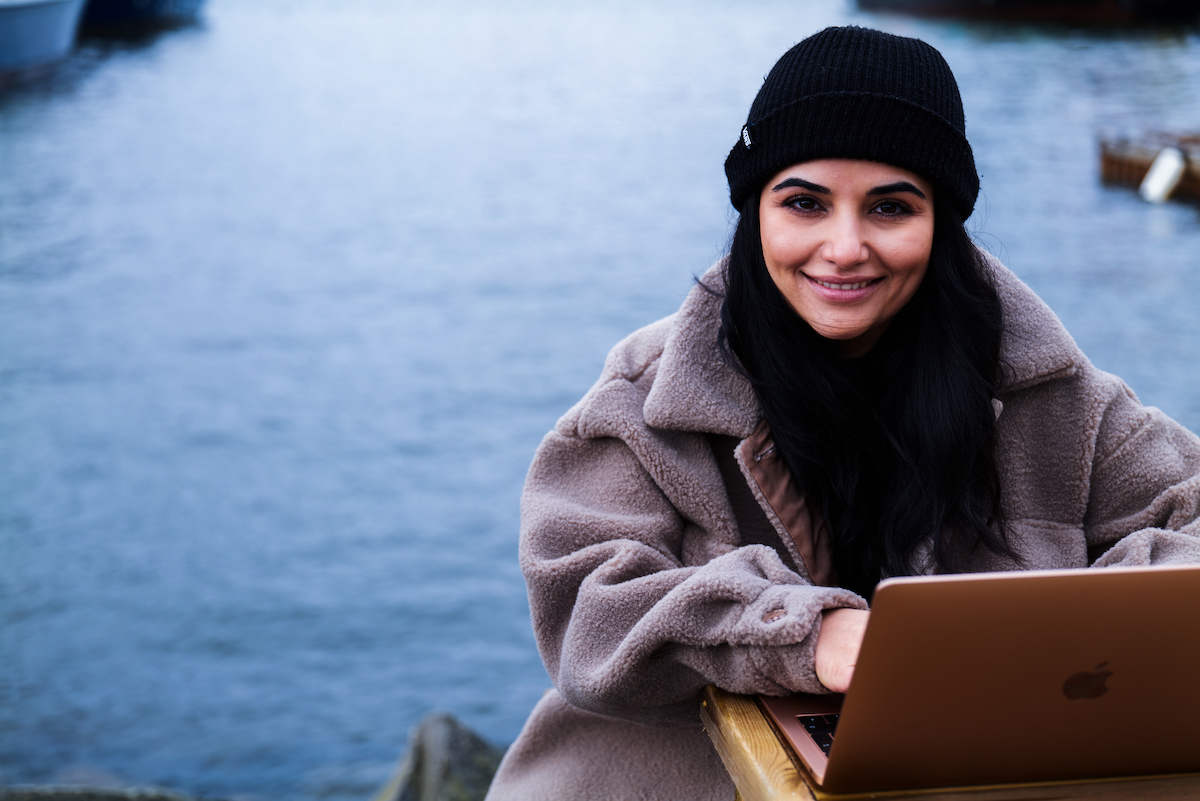 Bilde av ung kvinne i kåpe og lue, sitter utendørs og studerer på laptop foran havet.