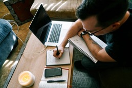 En mann sitter og skriver i notatboken sin samtidig som han sitter foran en datamaskin