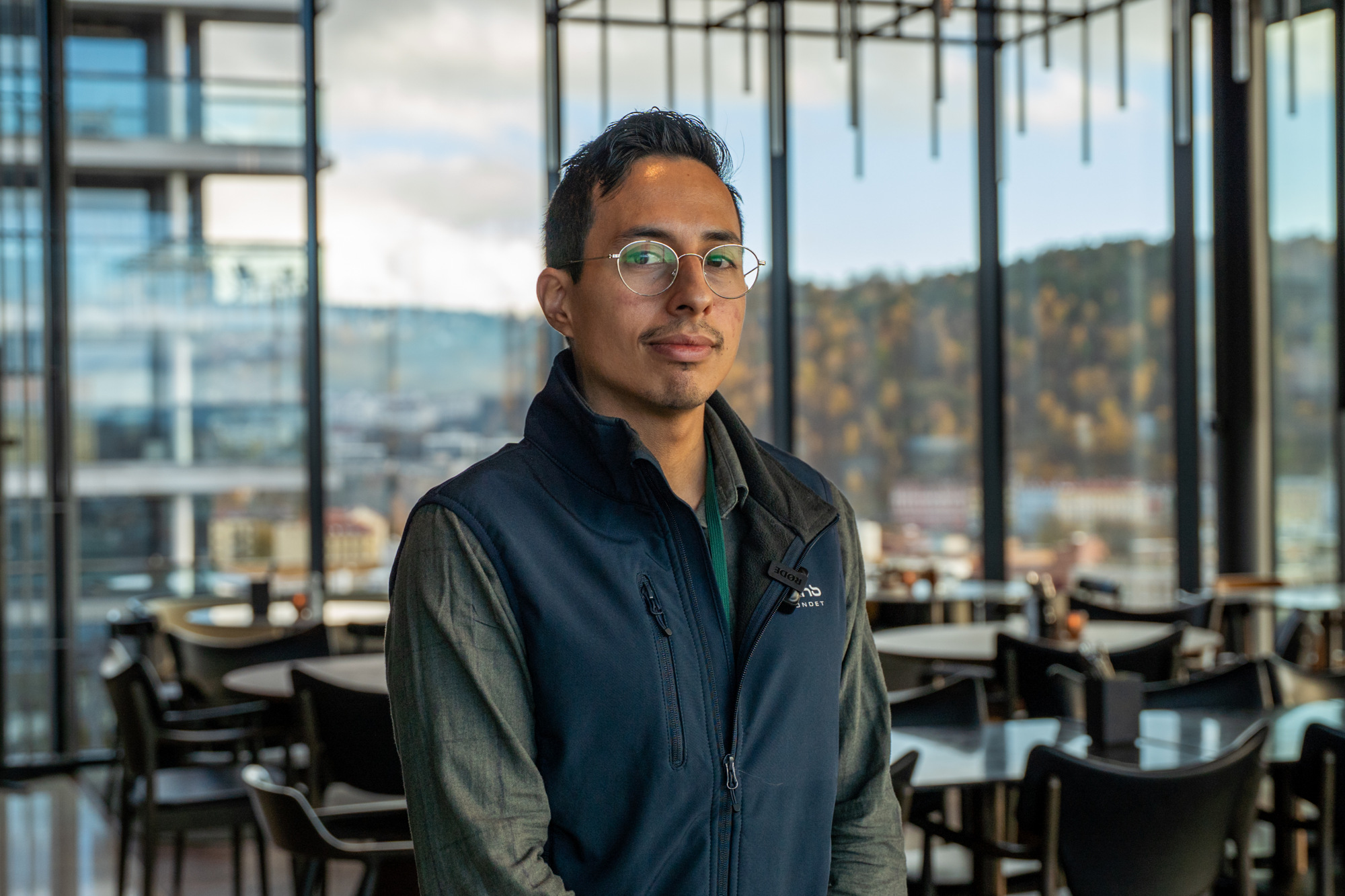 Luis står i en restaurant i øverste etasje, med en glassfasade bak seg som gir utsikt over byen.