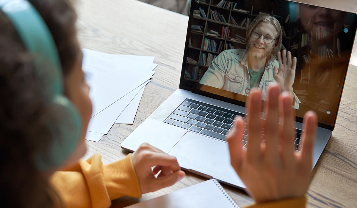 En kvinnelig student sitter ved et bord med en Mac fremfor seg, og har en videosamtale med en mann. De vinker til hverandre gjennom skjermen.  Kvinnen har blå hodetelefoner, en oransje genser og mørkt hår. Mannen på skjermen har blondt halvlangt hår, skjegg og briller.