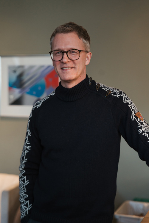 Portrett av Inge som står og smiler med en vegg og kunstverk i bakgrunnen.