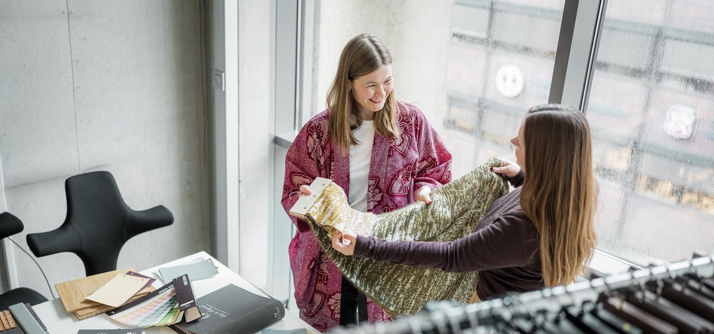 To studenter smiler til hverandre og holder opp tekstil i et lyst kontorlokale