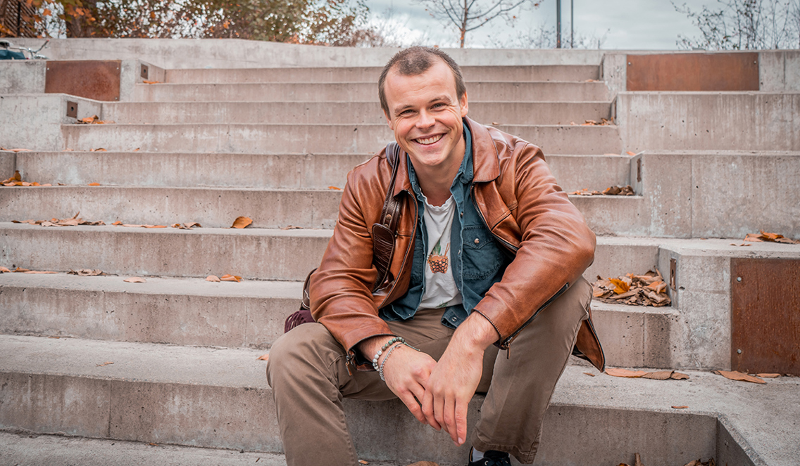 Peder Ulven sitter på en murtrapp utenfor campus Vulkan. Han har på seg en brun skinnjakke, har skinna hode og smiler pent til kamera. Han har armene lent over knærne.