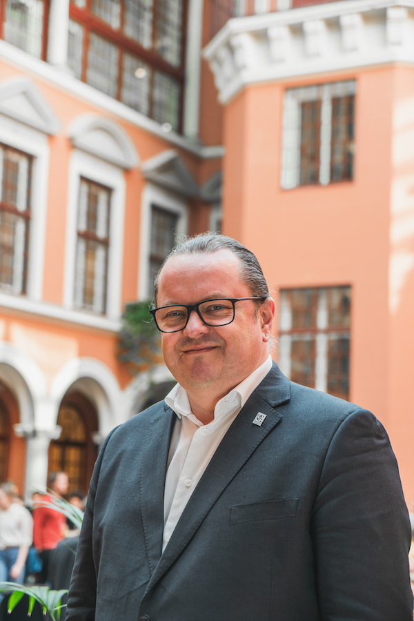 Arne H. Krumsvik ser inn i kamera på oransje bakgrunn. Han har på seg dress og briller.