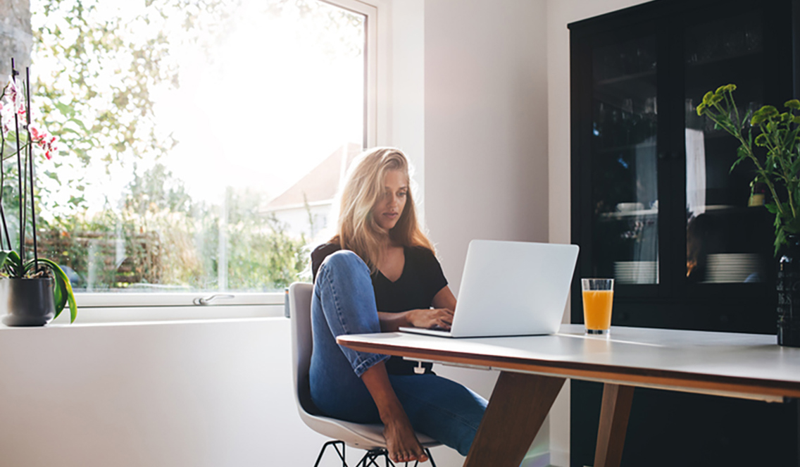 Jente som sitter ved frokostbordet med en laptop og et glass oransje leskedrikk. Bak henne er en hylle og et stort vindu som viser grønne omgivelser og solskinn.