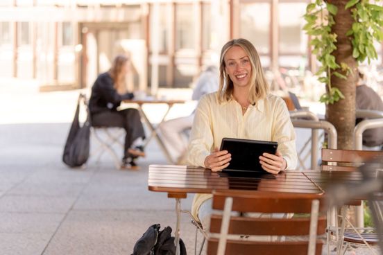 Person sitter ved et utendørs kafébord, holder et nettbrett og smiler inn i kamera