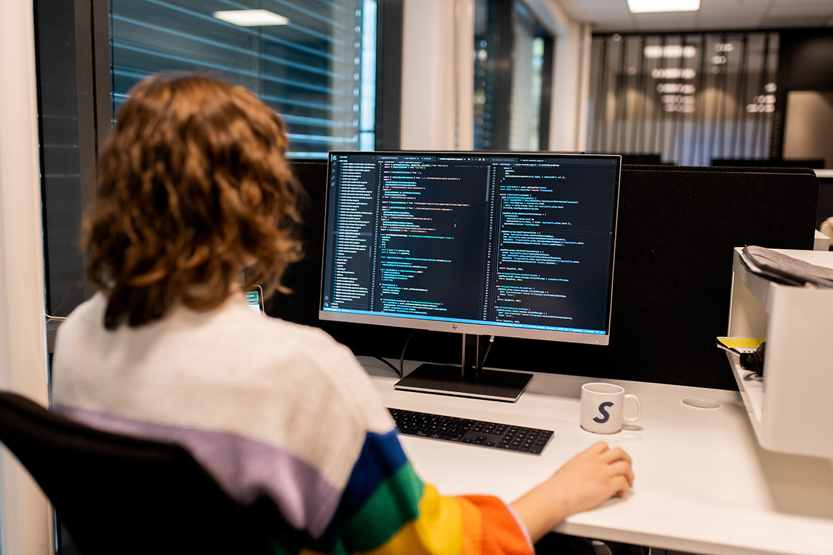 Foto av Stefanie Dziadkowiec som sitter ved en kontorpult og jobber på en datamaskin. På skjermen ser man mange linjer med koding.
