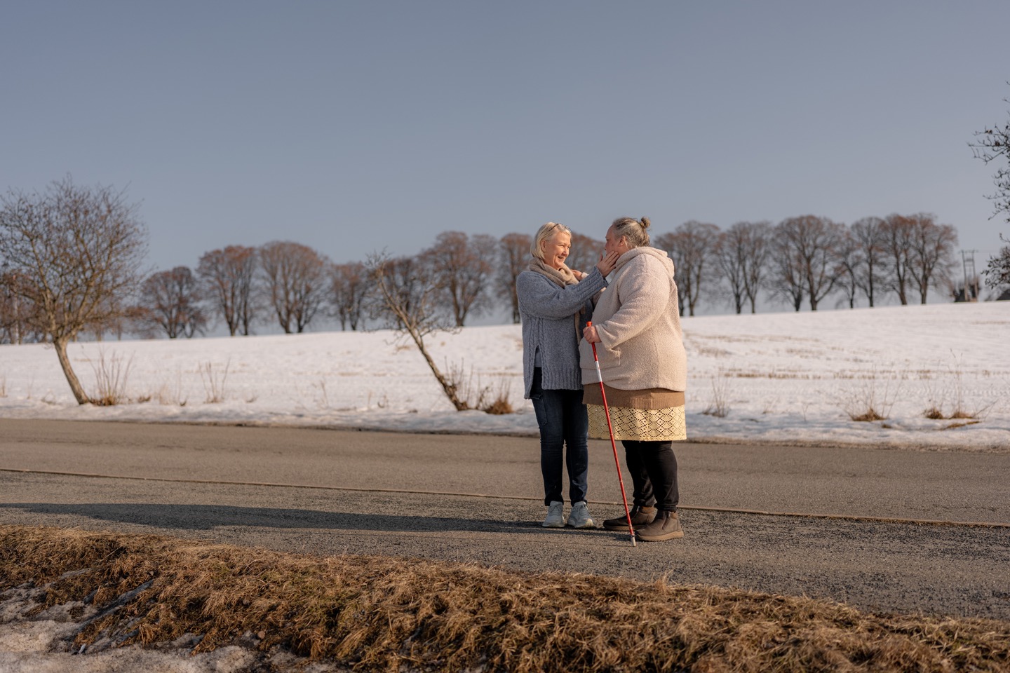 To personer står og snakker sammen på en landevei med snø i bakgrunnen