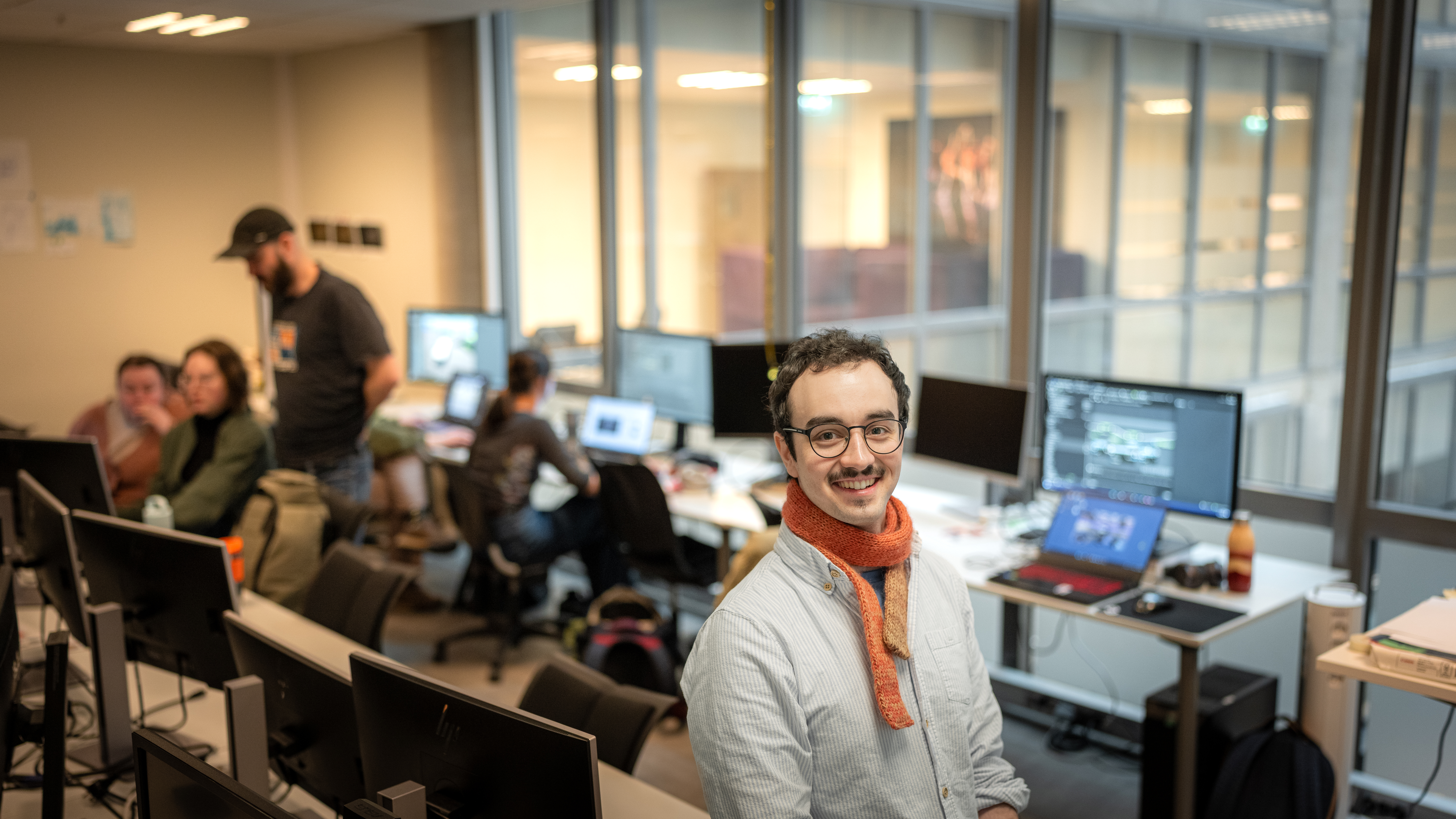 Student Shahin i undervisningsrom med datamaskiner, medstudenter og spillprosjekter i bakgrunnen.