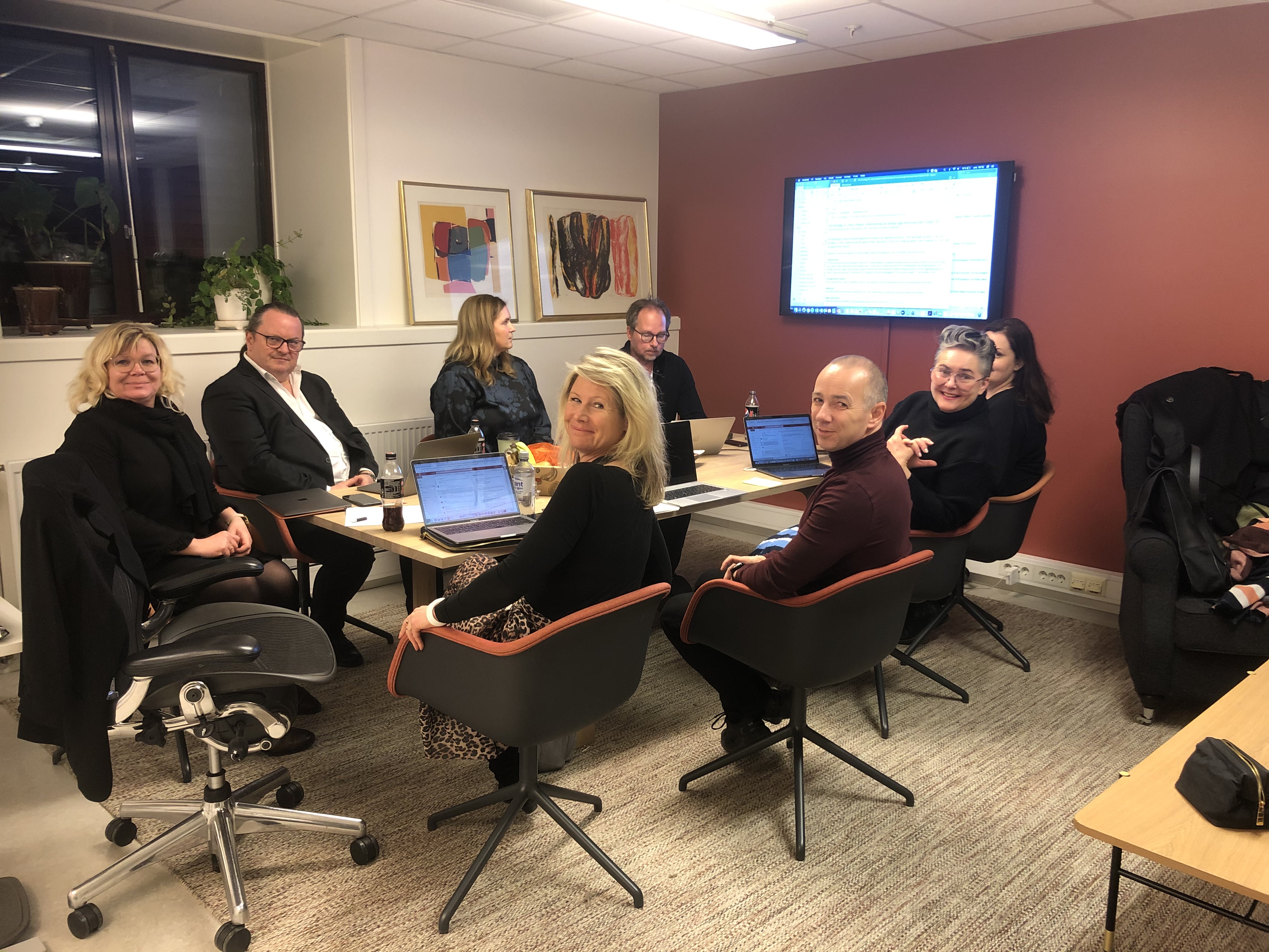 Åtte kvinner og menn sitter rundt et møtebord. Det er PC'er på bordet og de ser akkurat opp fra arbeidsoppgavene sine.