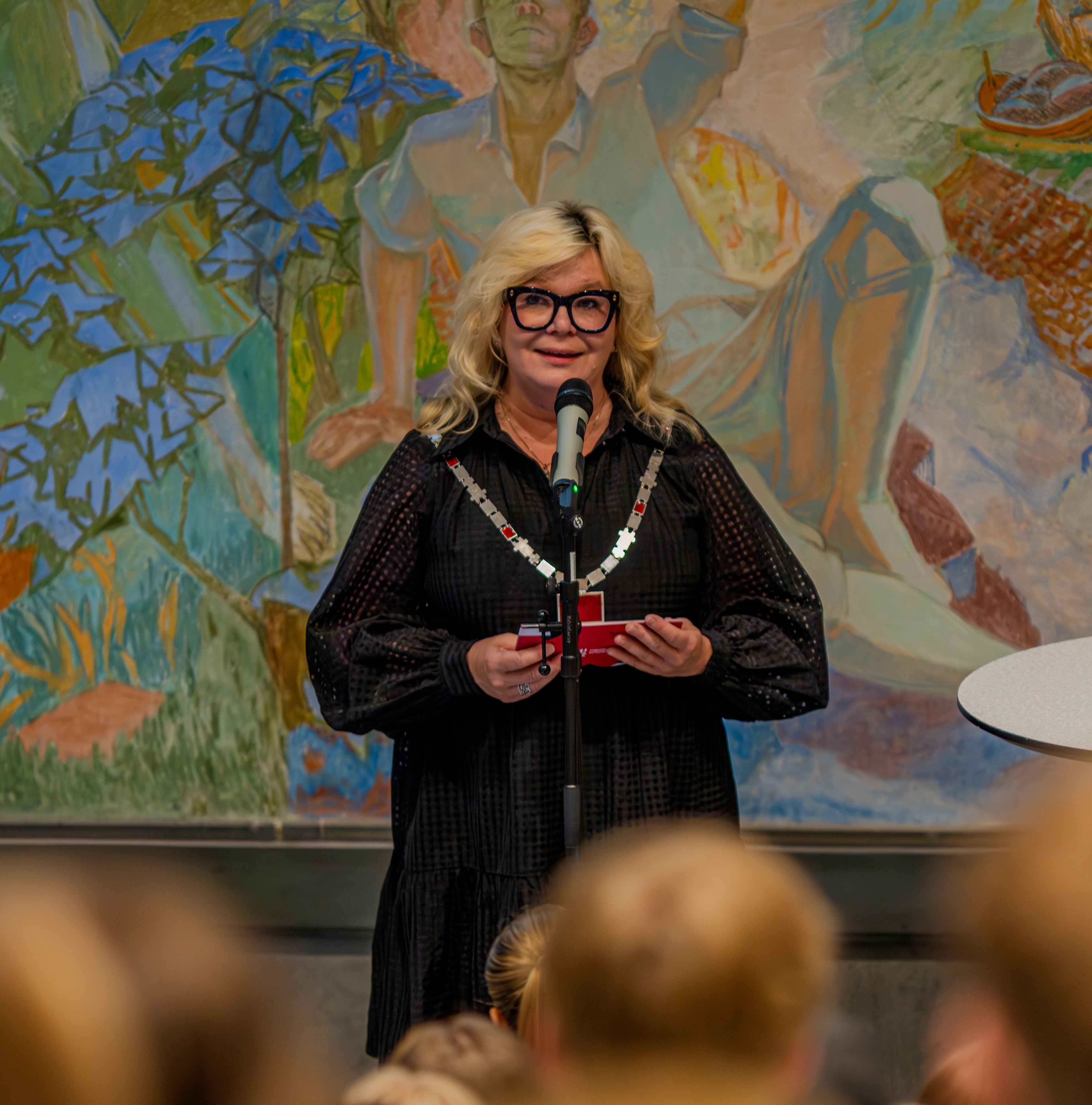 En pent kledd dame med briller og rektorkjede står foran en forsamling og snakker i en mikrofon. 