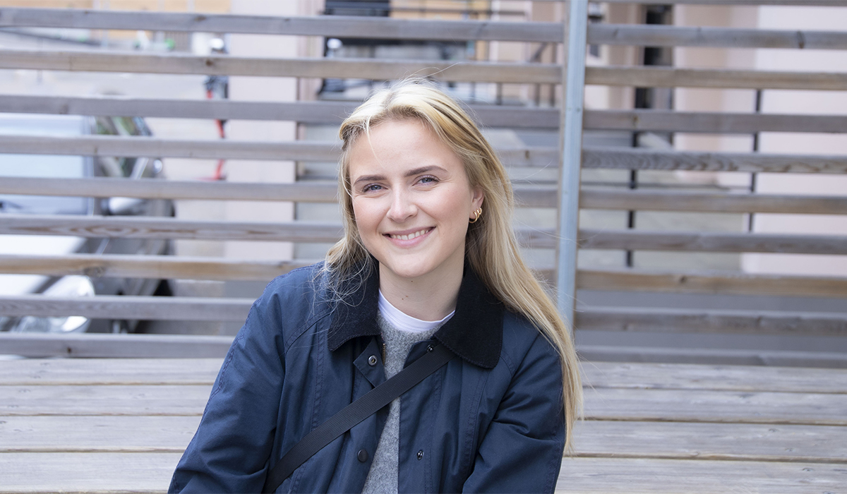 Kristiane Lunde sitter på en benk utenfor inngangen til Campus Fjerdingen med et brunt gjerde i bakgrunnen. Kristiane er blond og smiler. Hun har på seg en grå genser med en hvit topp under, øredobber, mørkeblå jakke og har en sort stropp over skulderen. 
