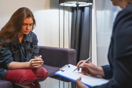 A young woman looks down on her hands while sitting in front of a doctor writing some notes