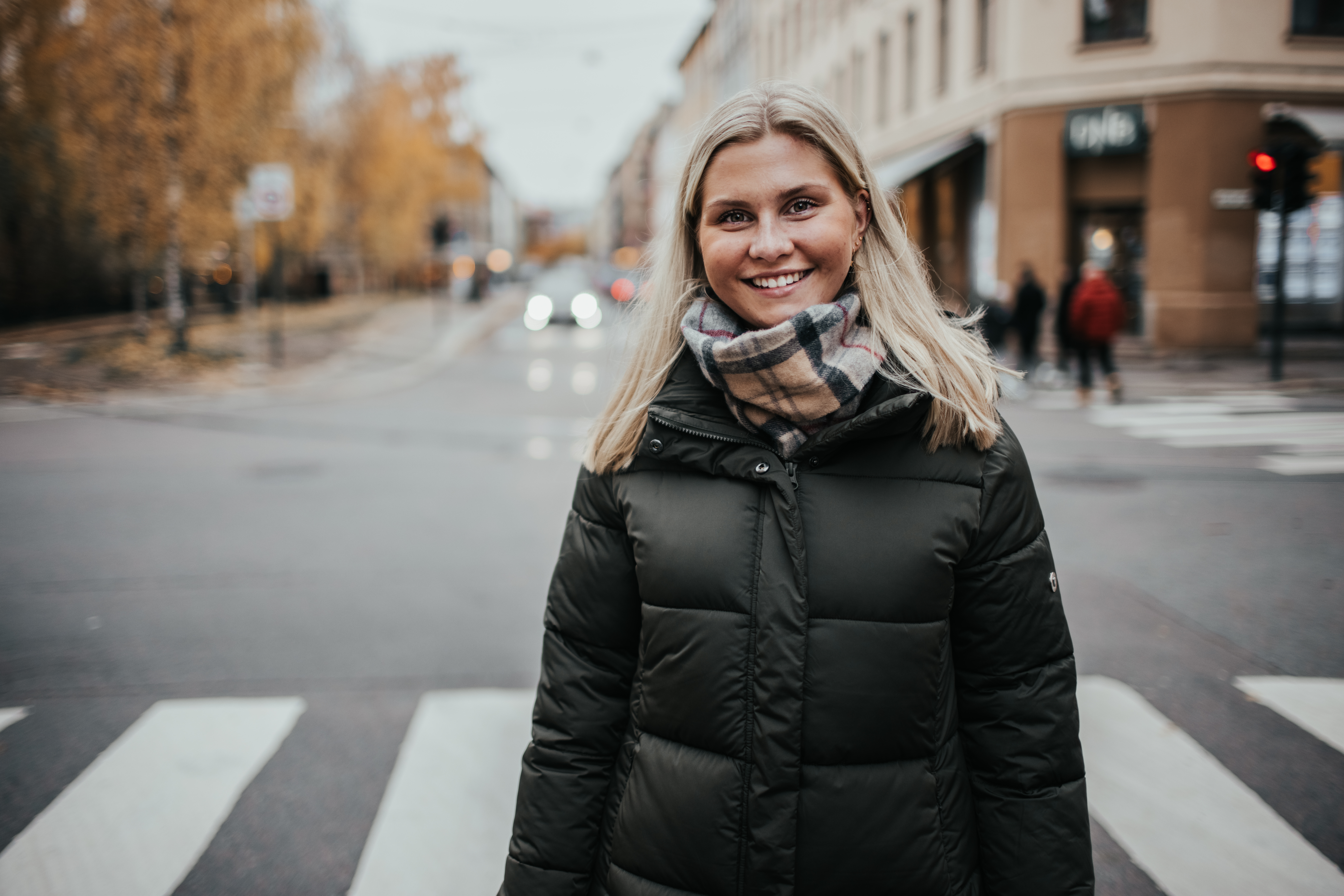 HR student Vilde står på fotgjengerfelt på Grünerløkka. Jente med mørk høstjakke, skjerf og blondt hår smiler.