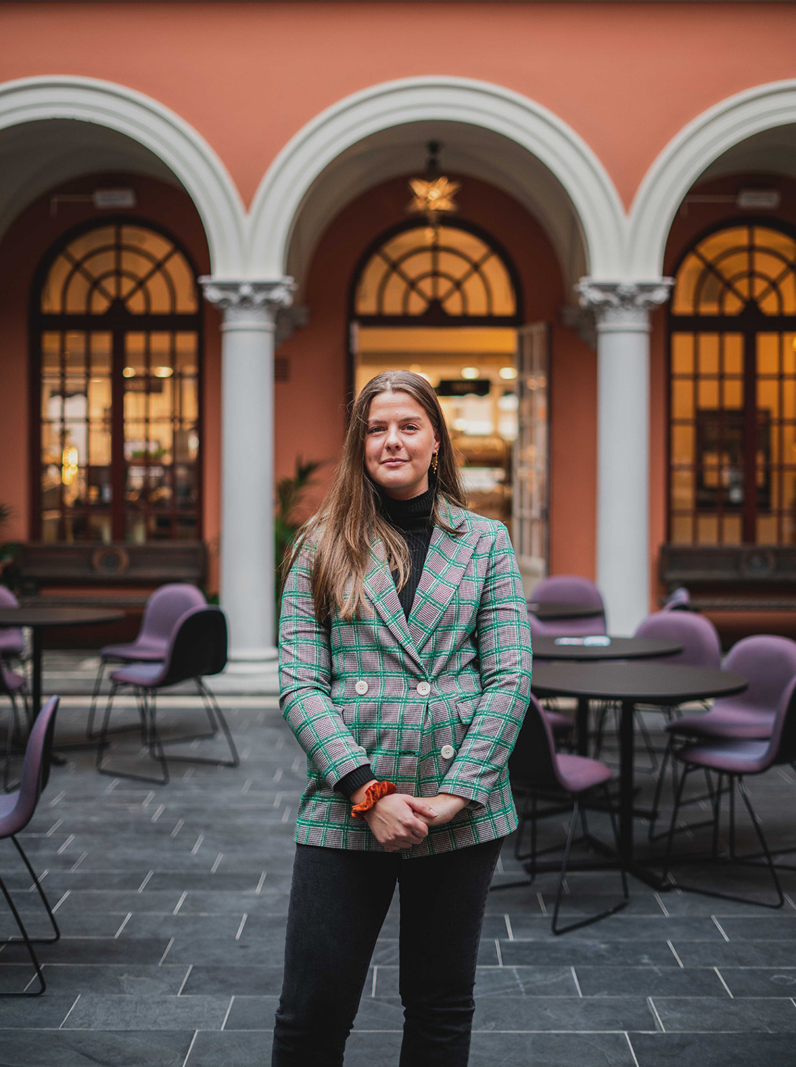 Caroline står i Ernst Hage på campus Kvadraturen. Hun har på seg en grønn blazerjakke med knapper og ruter. Jakken har grå trekk med grønne linjer. Hun har lagt brunt hår og har hendene foldet foran magen. 
