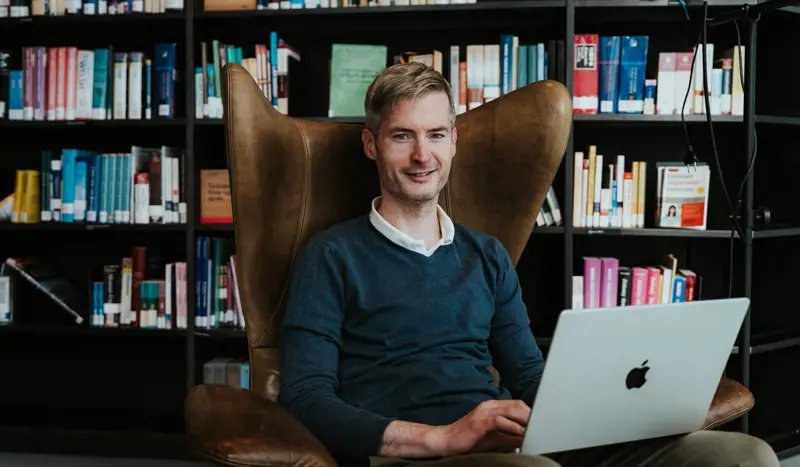 Mann sitter i en stol på et bibliotek og studerer på en mac.