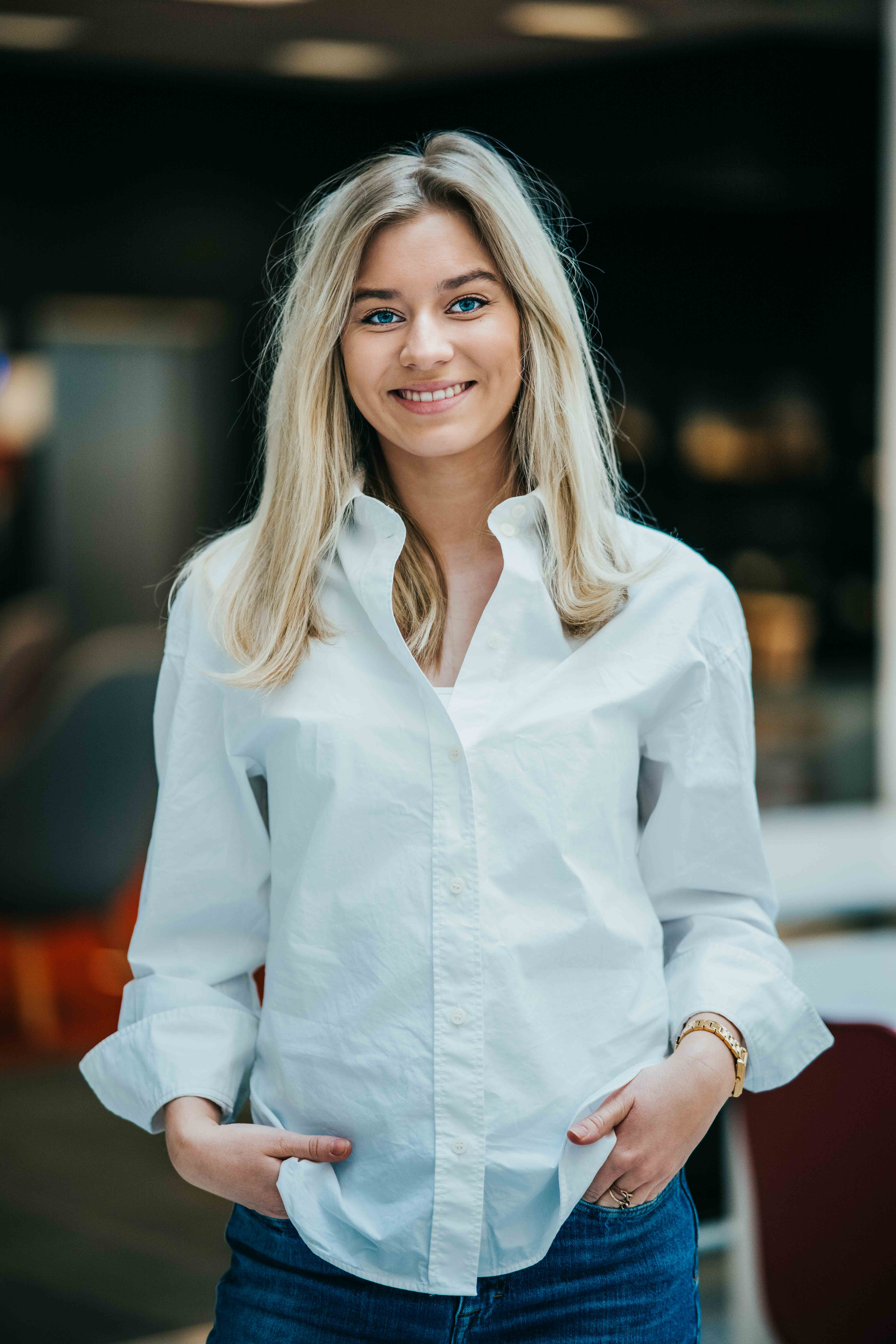 Ung, blond dame i hvit topp og blå jeans smiler til kamera.
