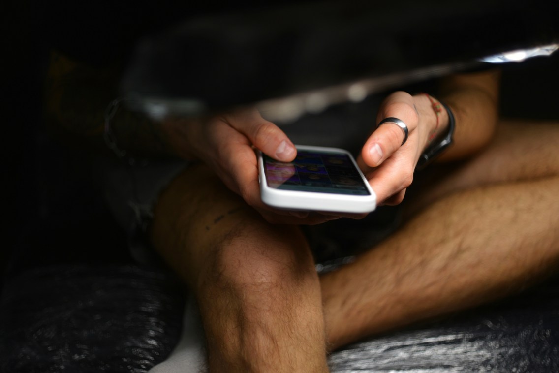 Ung manns hender og ben mens han sitter i mørket og ser på mobilskjermen