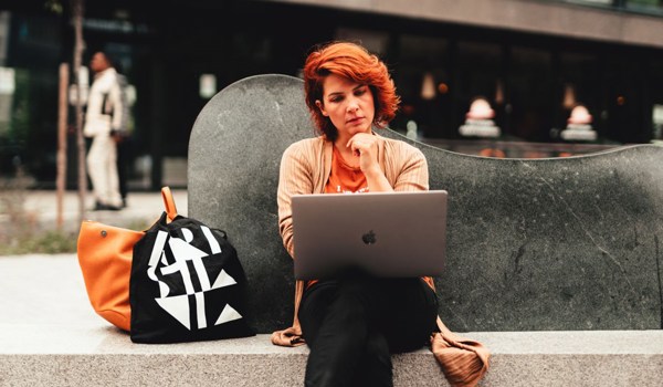 Foto av kvinne som sitter på en benk utendørs med en laptop på fanget.