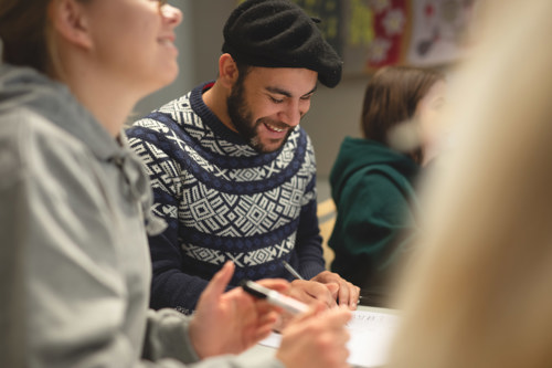 En student sitter mellom to andre studenter, han ser ned og smiler.