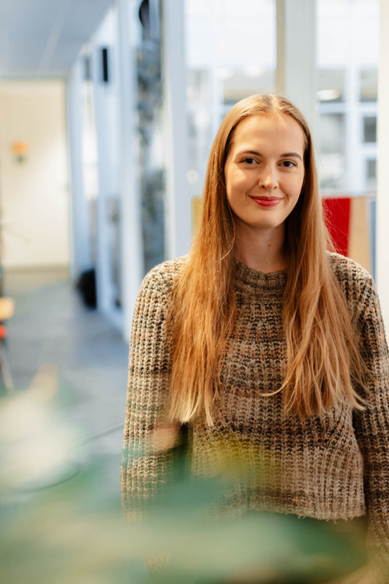 Benedicte står smilende i et rom bak en plante 