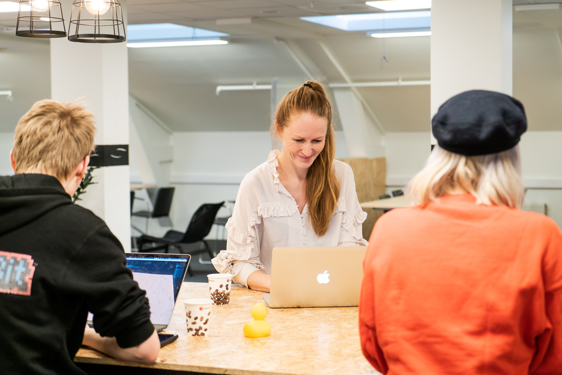 Kvinnelig underviser og to studenter sitter rundt et bord med hver sin datamaskin.