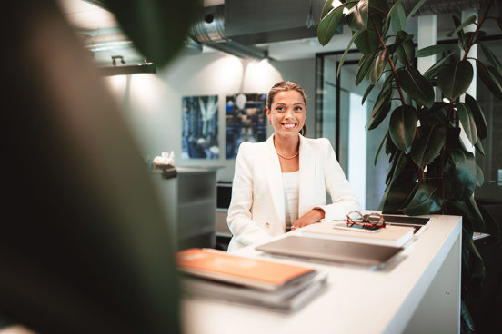 Bilde av ung kvinne i penklær som sitter mellom planter på et kontor og smiler til kamera 