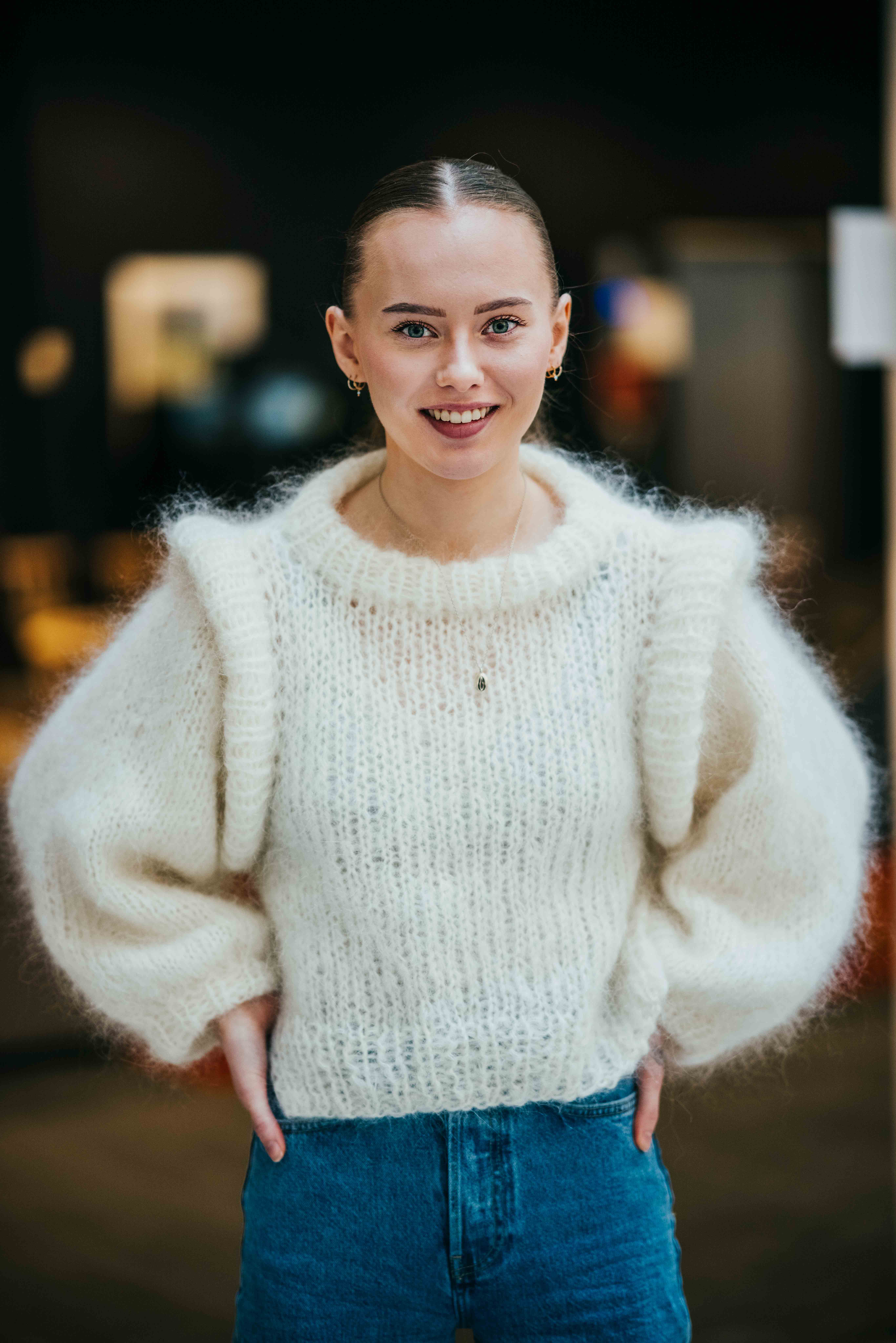 Ung kvinne i hvit genser og blå jeans smiler til kamera.