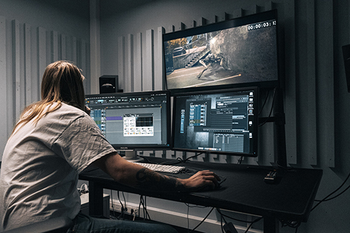 Student i studio som jobber på miksepult og PC med tre skjermer.