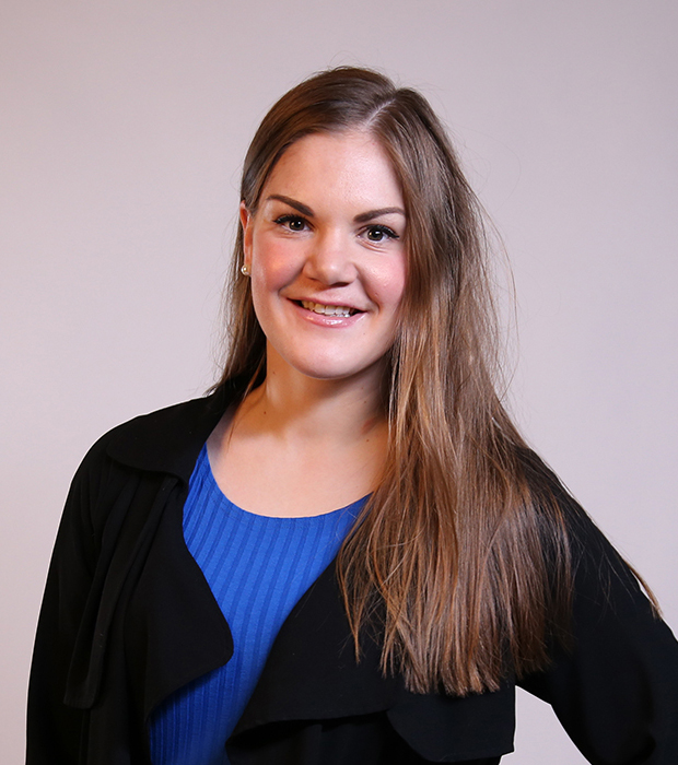 Portrettbilde av Ingrid. Hun har den ene hånden plassert på hoften. Hun har på seg en blå t-skjorte med en sort cardigan over. Hun har langt brunt hår som er lagt over den ene skulderen.