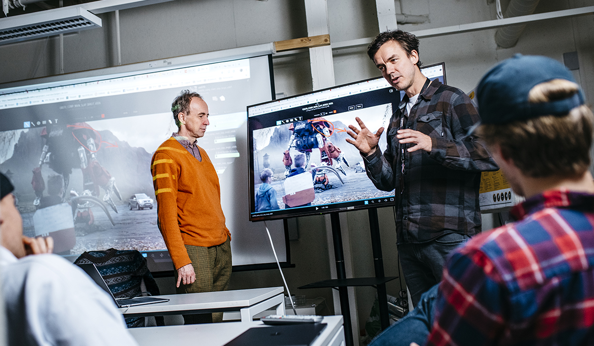 Hans Gunnar og Morten Moen står foran i et klasserom. Mellom dem kan man se en tv-skjerm med et teknolgisk program oppe. På skjermen er det ulike figurer og karakterer. 