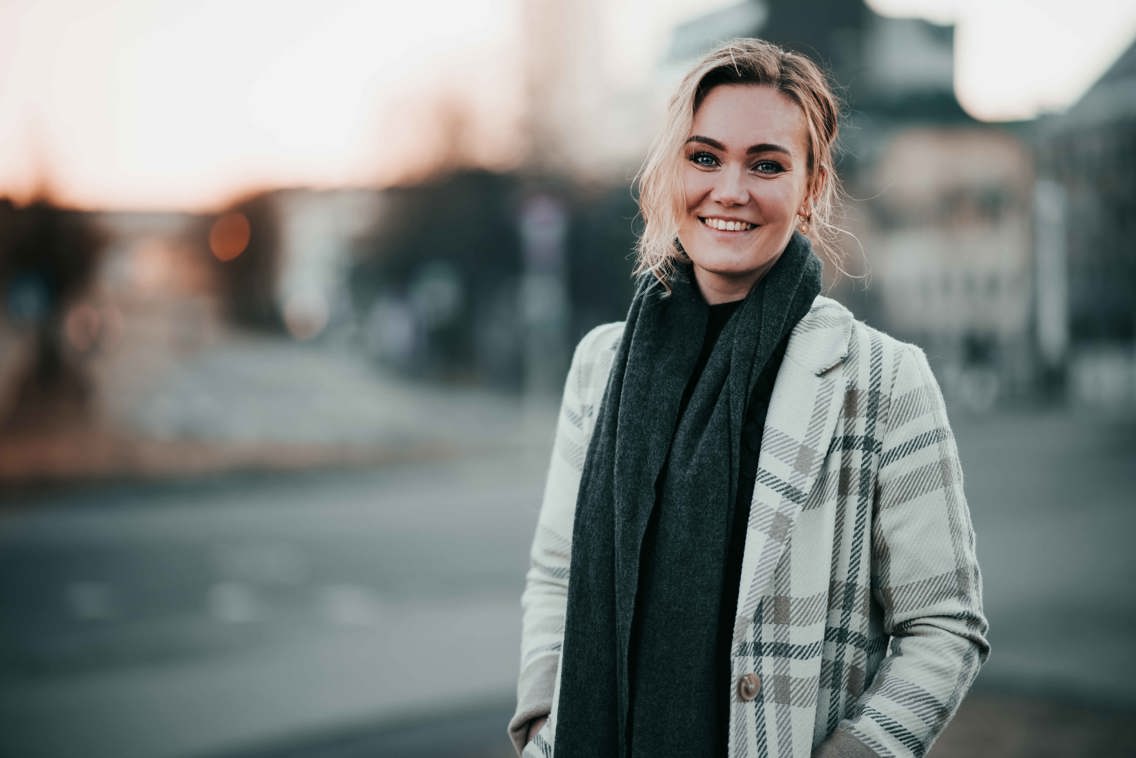 Ung kvinne i lys kåpe og mørkt skjerf smiler til kamera
