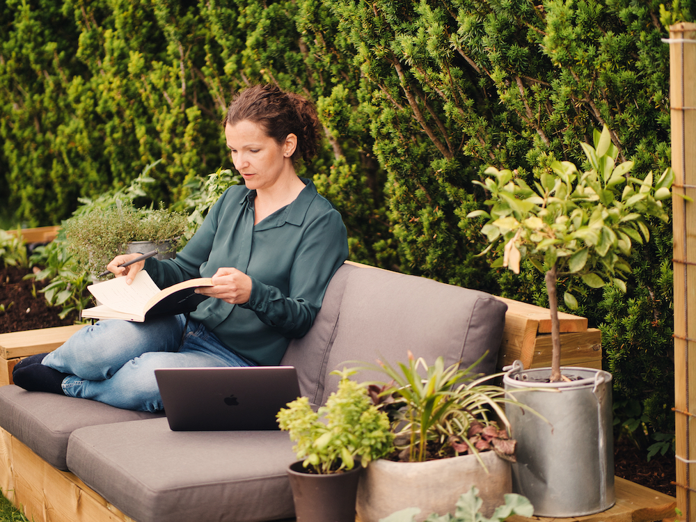 Kvinne som sitter i hagen. Hun leser i en bok og har en laptop ved siden av seg. 