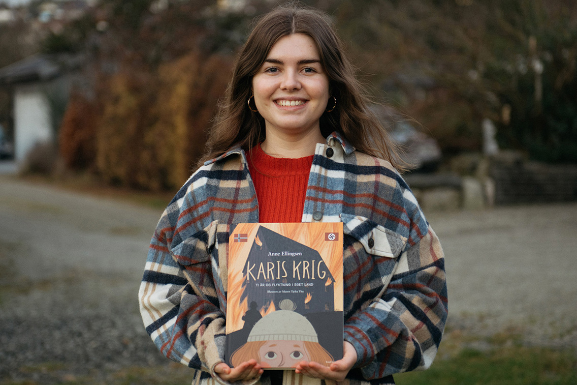 Maren Tjelta Thu har mørkt, langt hår og har på seg en rød genser og en rutete jakke. Hun holder boken Karis krig opp foran seg, og smiler mot kamera.