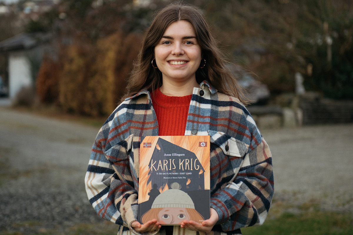 Maren Tjelta Thu har mørkt, langt hår og har på seg en rød genser og en rutete jakke. Hun holder boken Karis krig opp foran seg, og smiler mot kamera.