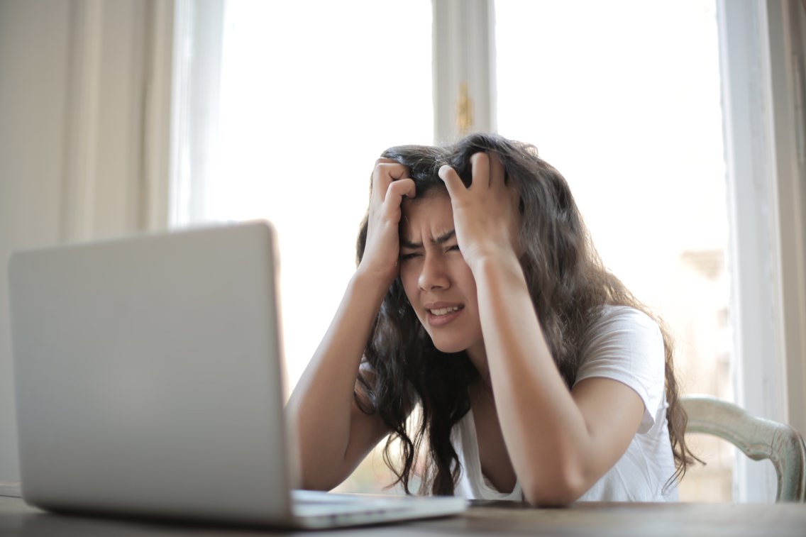 Frustrert kvinne tar seg til hodet mens hun sitter foran en laptop.