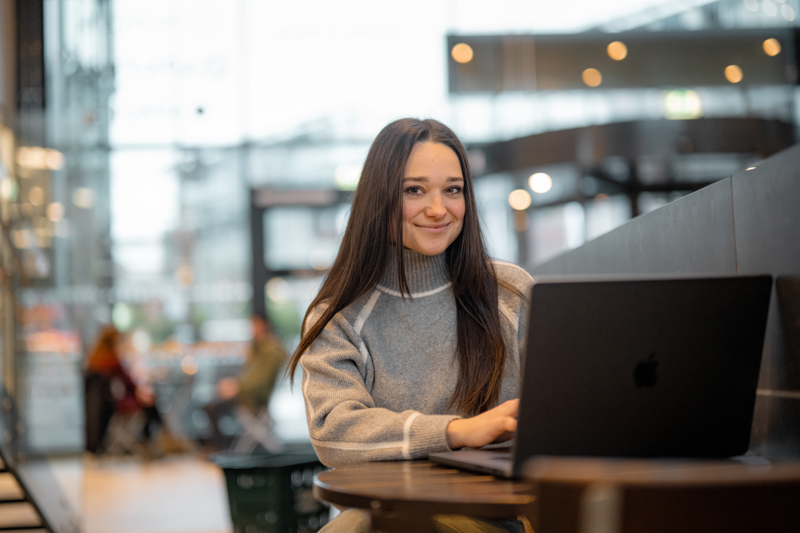 Student jobber på laptop i åpent fellesområde med store vinduer og sitteplasser