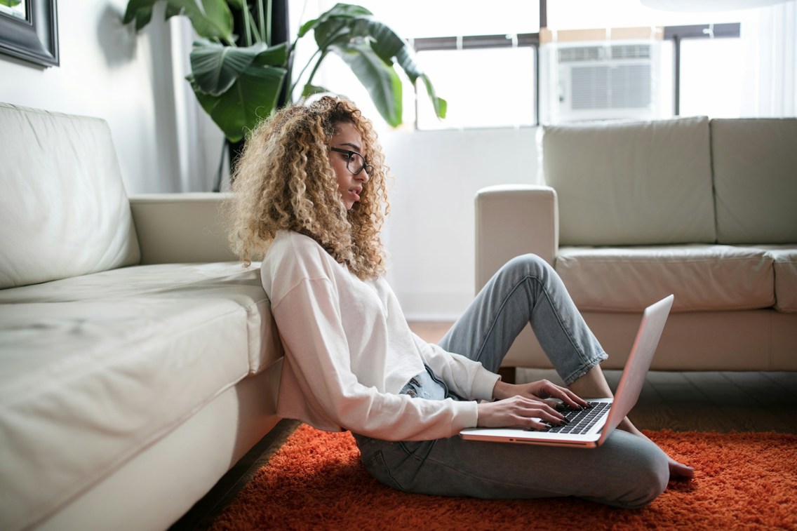 Kvinne sitter på gulvet med laptop og ryggen mot sofaen