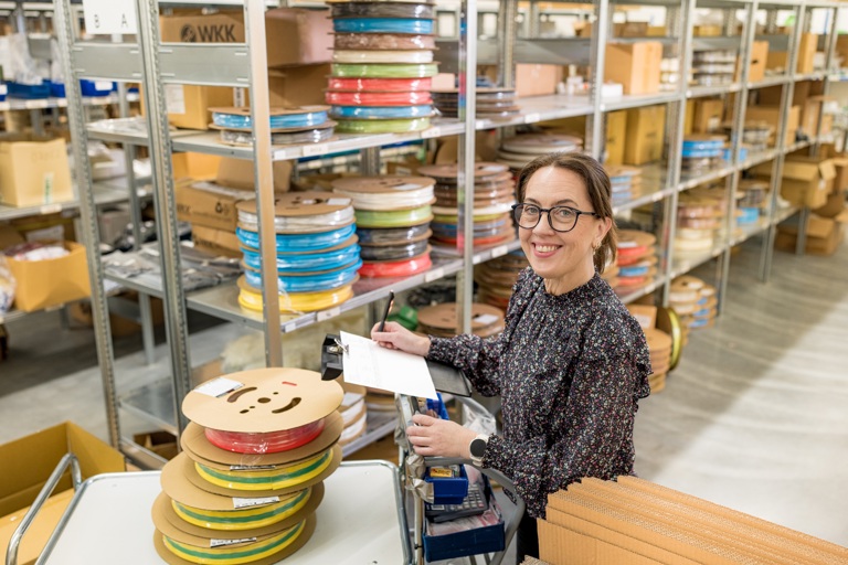 Logistikkarbeider smiler inn i kamera foran hyller med varer