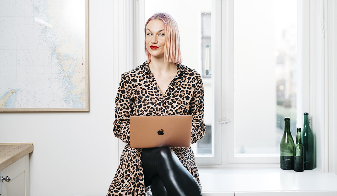 Christina Fraas sitter på kontoret sitt med en laptop i fanget. Hun har på seg sorte skinnbukser og en leoparttopp. Bak henne er et vindu og det er bilder på veggene. Hun ser rett inn i kameraet med et bestemt selvsikkert uttrykk i ansiktet. Blondt hår og rød leppestift. 