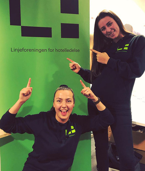 To kvinnelige studenter poserer smilende foran en grønn roll-up med logoen til Linjeforeningen for hotelledelse. 